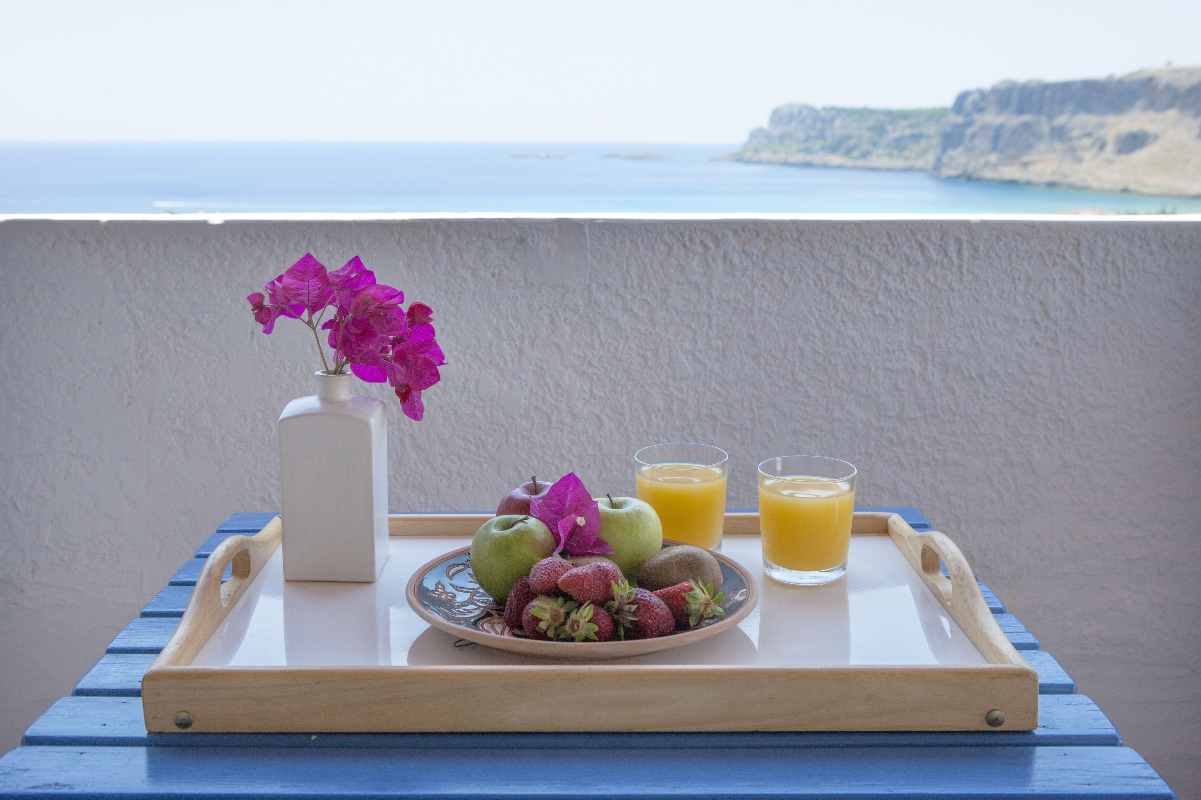 Una bandeja con frutas, un jarrón con flores y vasos de jugo de naranja en un balcón con vista al mar.