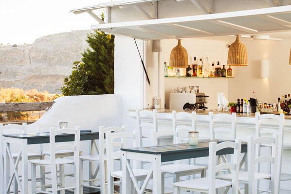 An outdoor bar area with white chairs and tables, a bar counter with bottles, and a view of a rocky landscape.