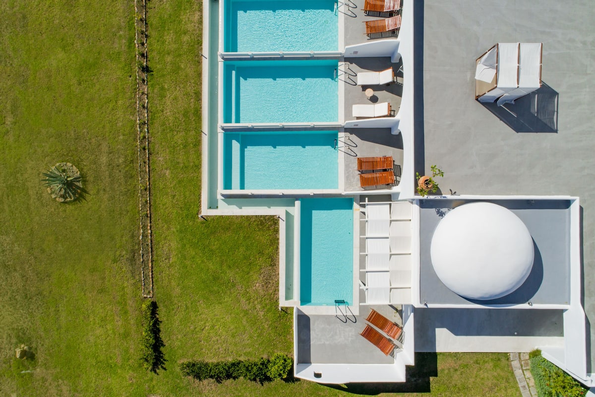 An aerial view of a hotel with three swimming pools, a large spherical water feature, and a grassy area with a palm tree.