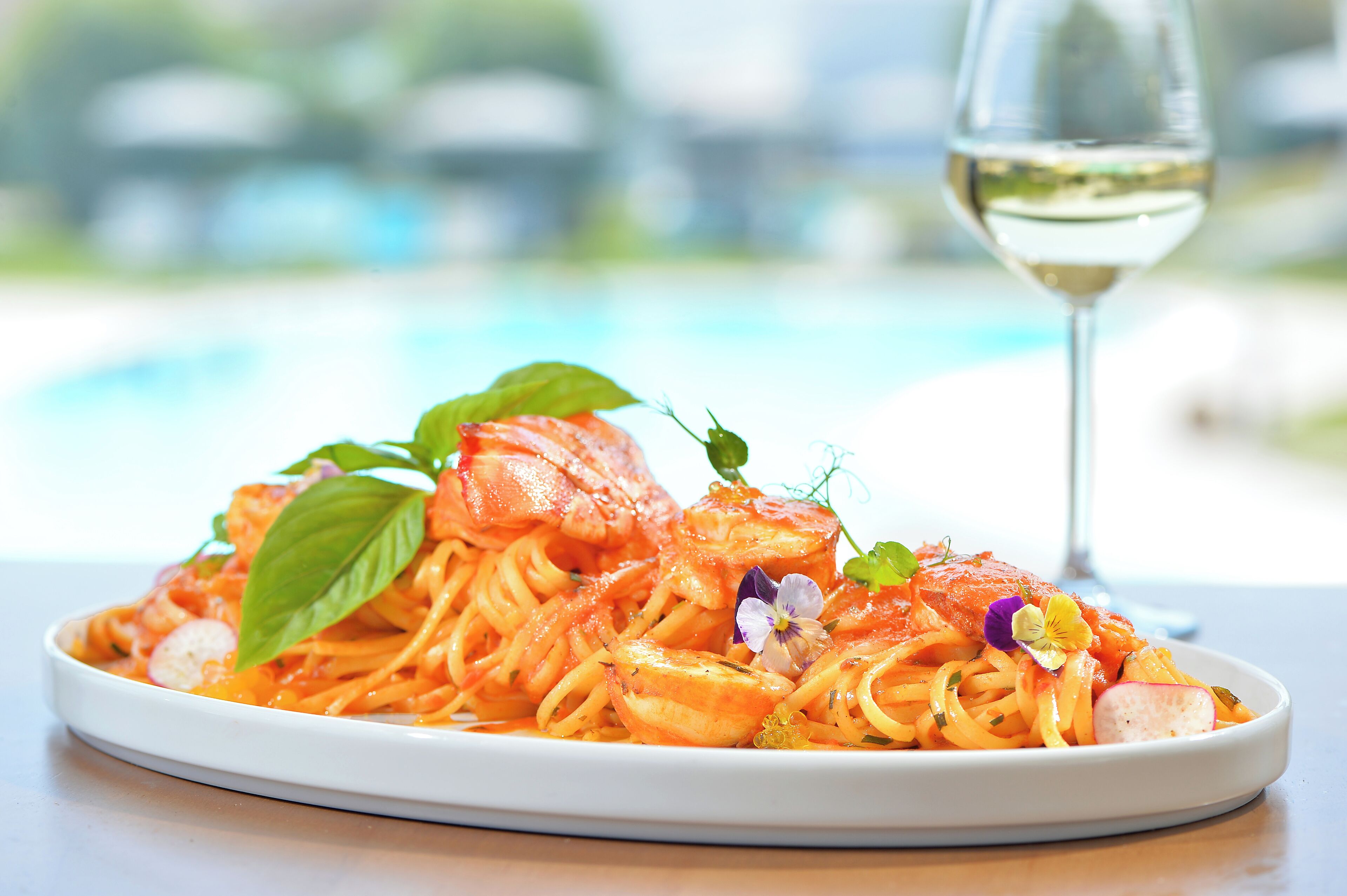 A plate of spaghetti with tomato sauce, garnished with basil and flowers, and a glass of white wine on a table with a blurred pool in the background.