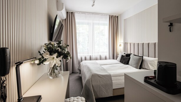 A modern hotel room with a large bed, a bedside table, a flat-screen TV, a vase with flowers, and a window with curtains. - Hotel Delta (Kraków)