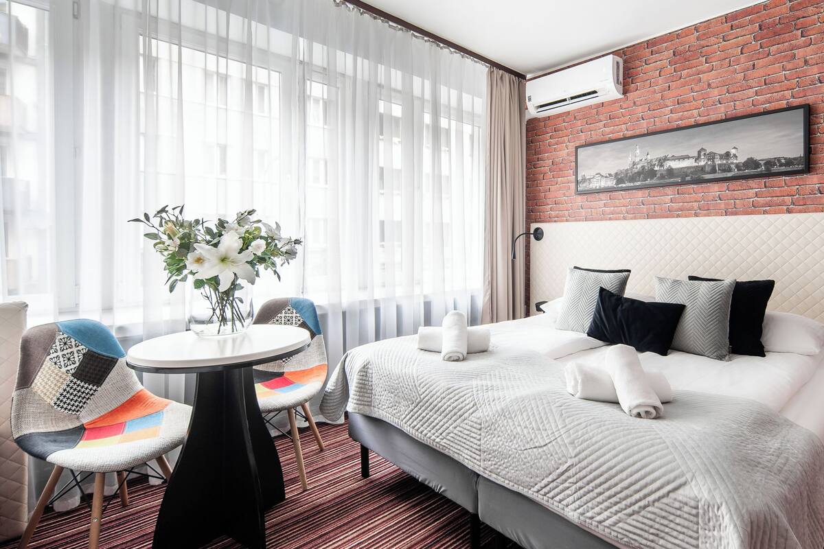 Une chambre d’hôtel avec un grand lit, une petite table ronde ornée d’un vase de fleurs, une chaise avec un coussin coloré et un mur de briques sur lequel est accroché un tableau.
