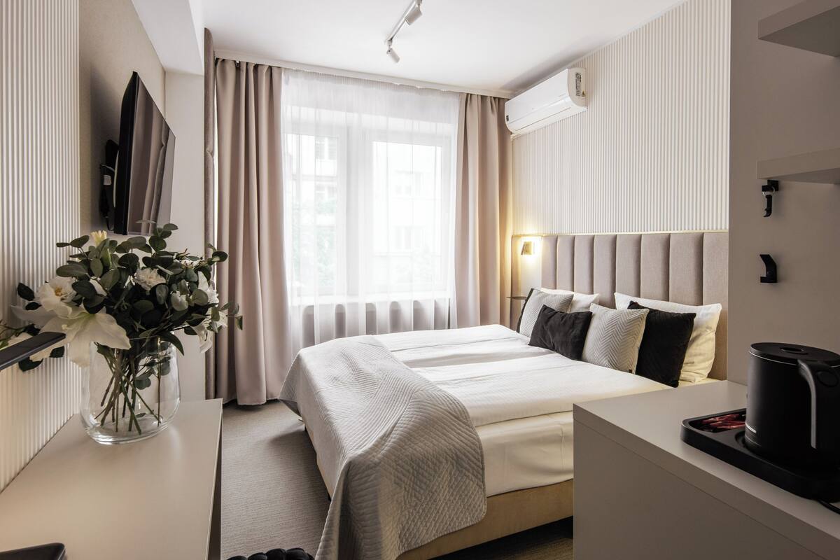 Une chambre d’hôtel comprenant un lit, une table de chevet, une lampe, un appareil de climatisation et un vase avec des fleurs.