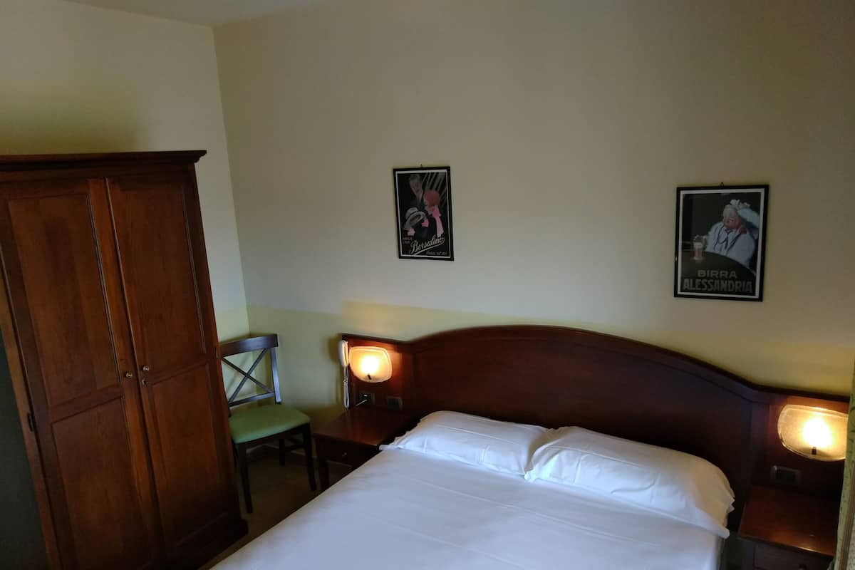 Une chambre d’hôtel avec une grande armoire en bois, un lit simple avec du linge de lit blanc, deux tables de chevet avec des lampes et deux tableaux encadrés au mur.