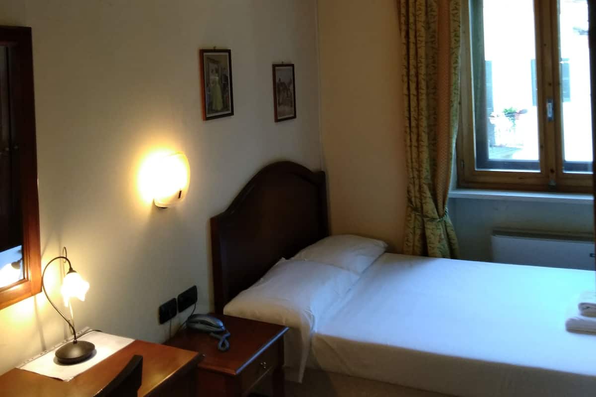 Une chambre d’hôtel avec un lit simple, un bureau avec une lampe, un téléphone et une fenêtre avec des rideaux.