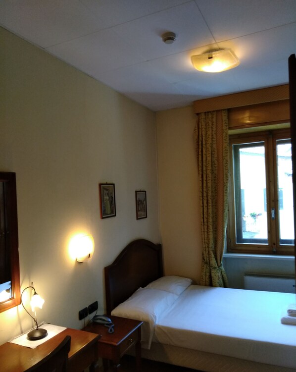 A hotel room with a single bed, a desk with a lamp, a telephone, and a window with curtains. - Hotel Belvedere (Alice Bel Colle)