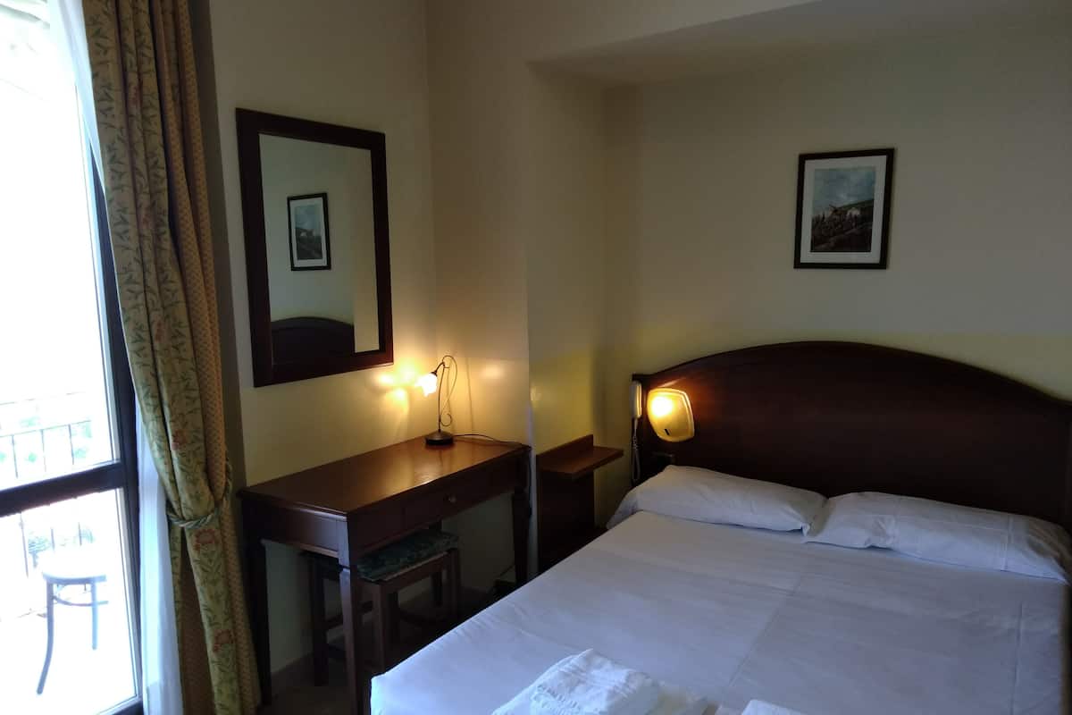 Une chambre d’hôtel avec un lit, un bureau, une chaise, une lampe et une fenêtre avec des rideaux.