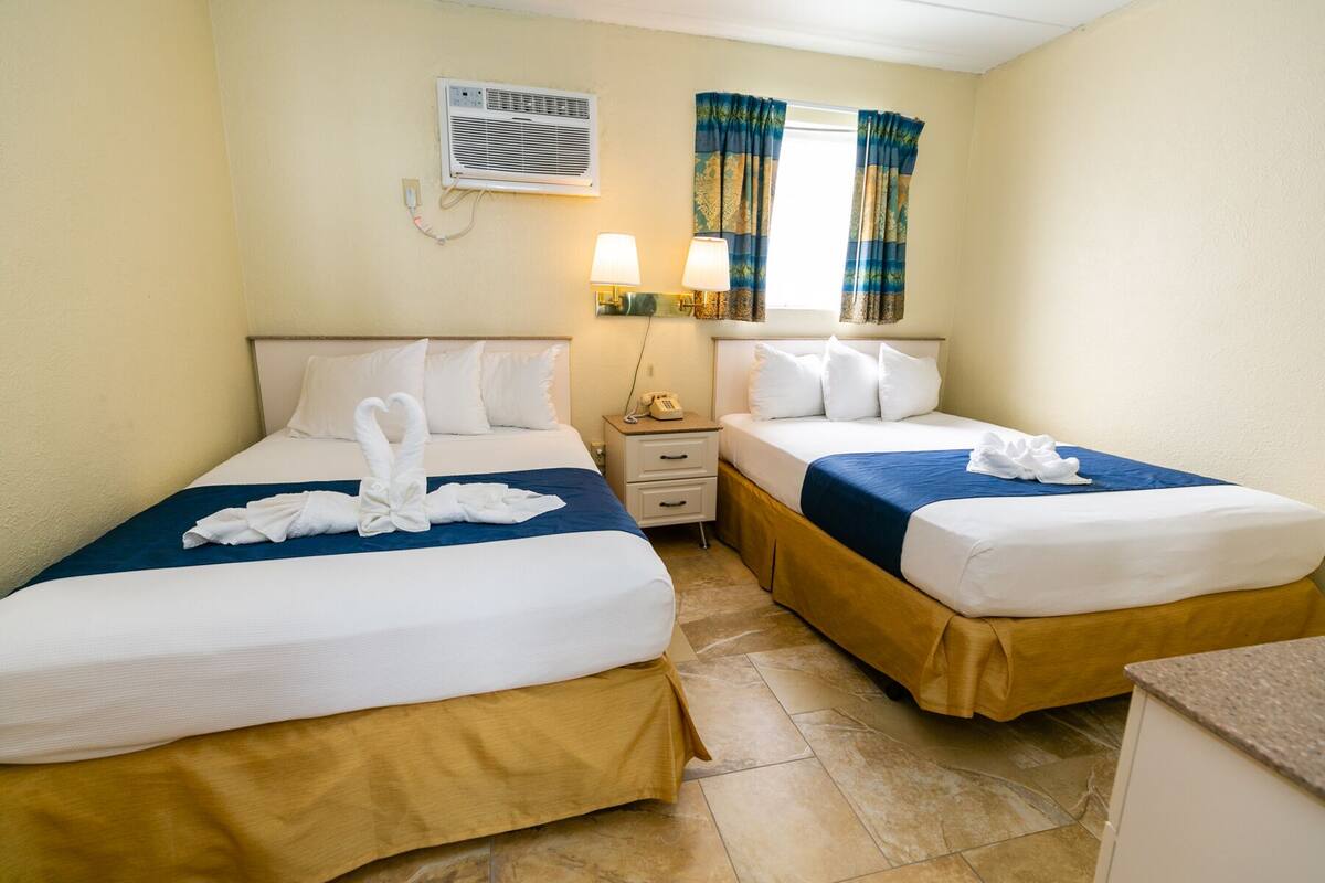 A hotel room with two beds, each with white linens and towels arranged in a heart shape. There is a bedside table with a lamp and a telephone. The room has a window with blue curtains and an air conditioning unit mounted on the wall.