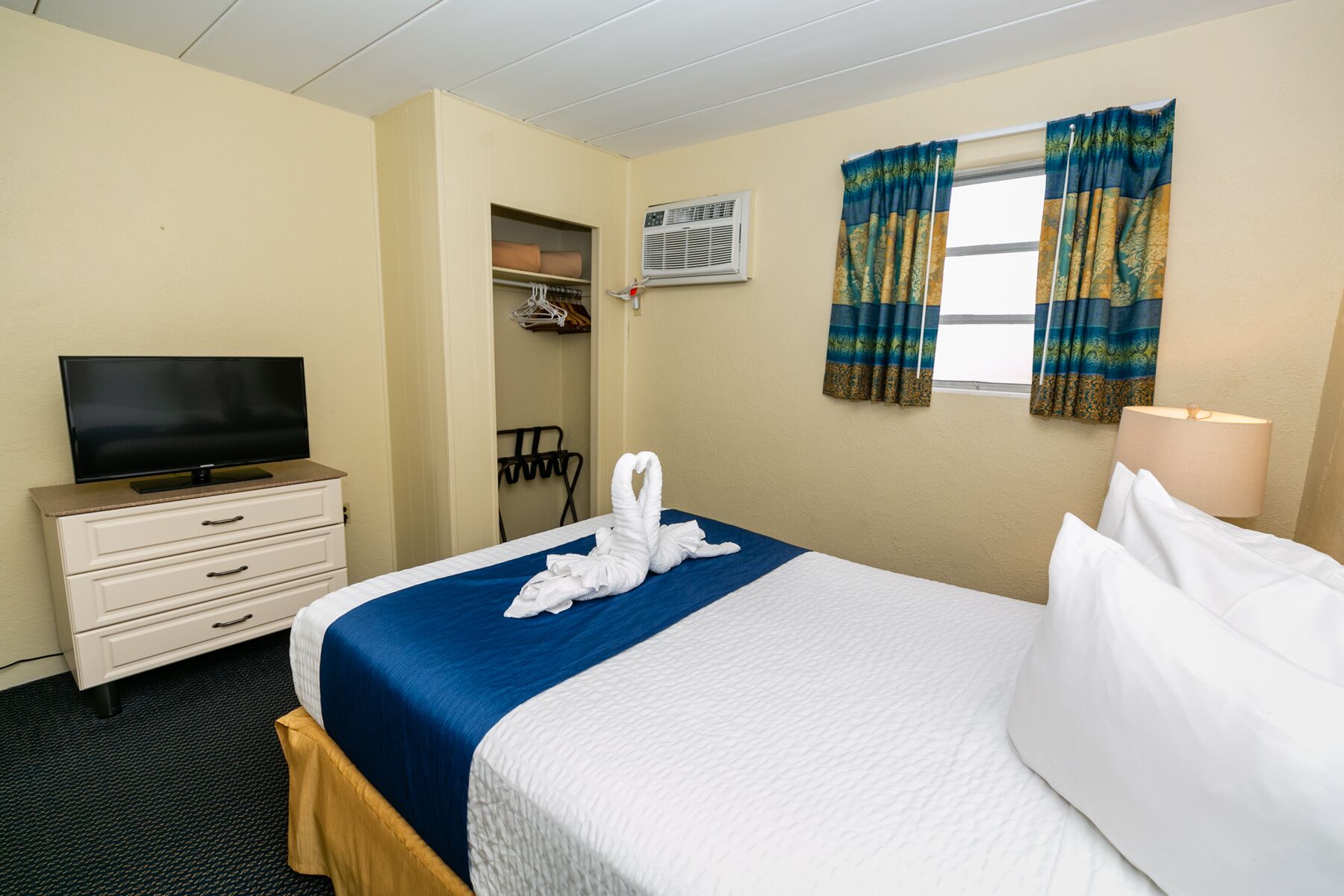 A hotel room with a bed, a television on a dresser, a window with curtains, and a coat rack.