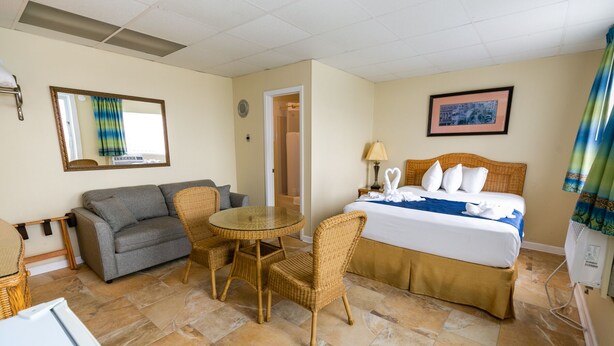 A hotel room with a bed, a gray sofa, a wicker dining table, and a small refrigerator.