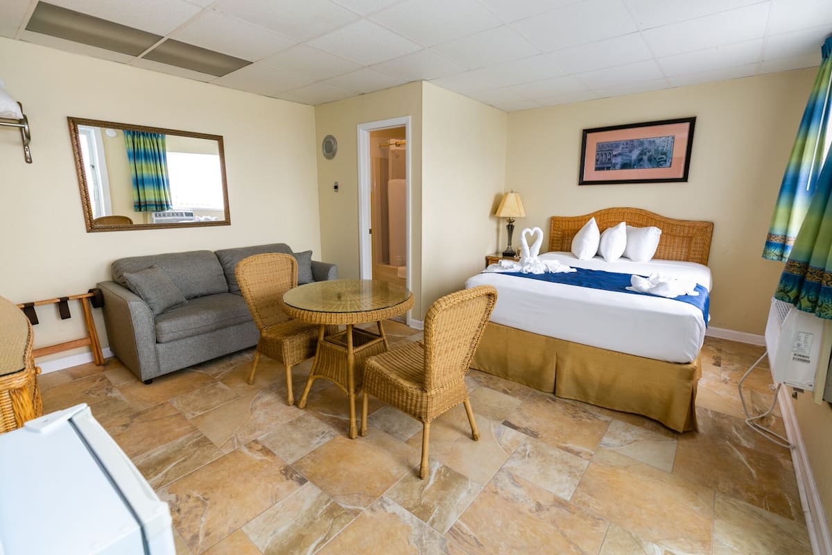 A hotel room with a bed, a gray sofa, a wicker dining table, and a small refrigerator.