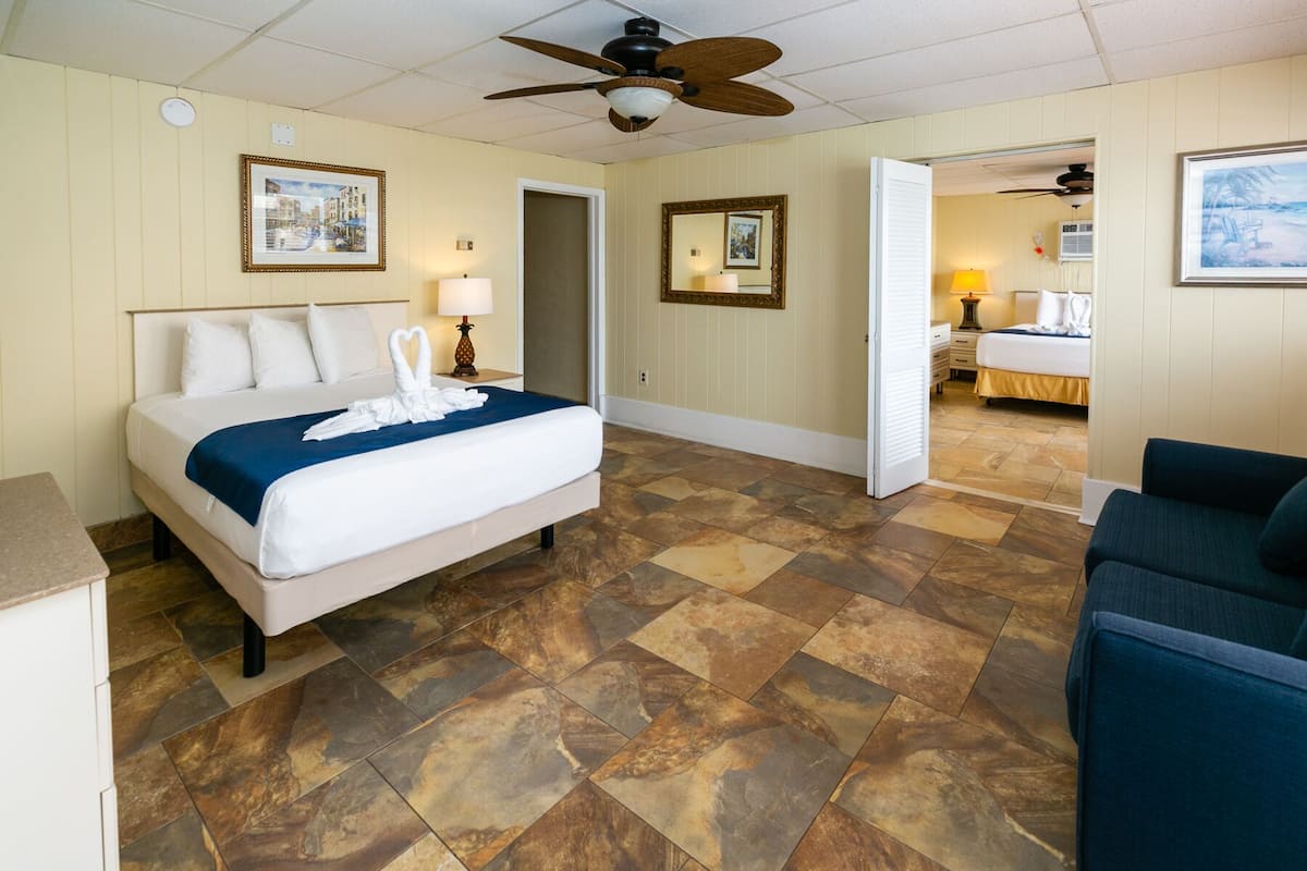 A hotel room with a large bed, a ceiling fan, a bathroom with a shower, and a view of the beach.