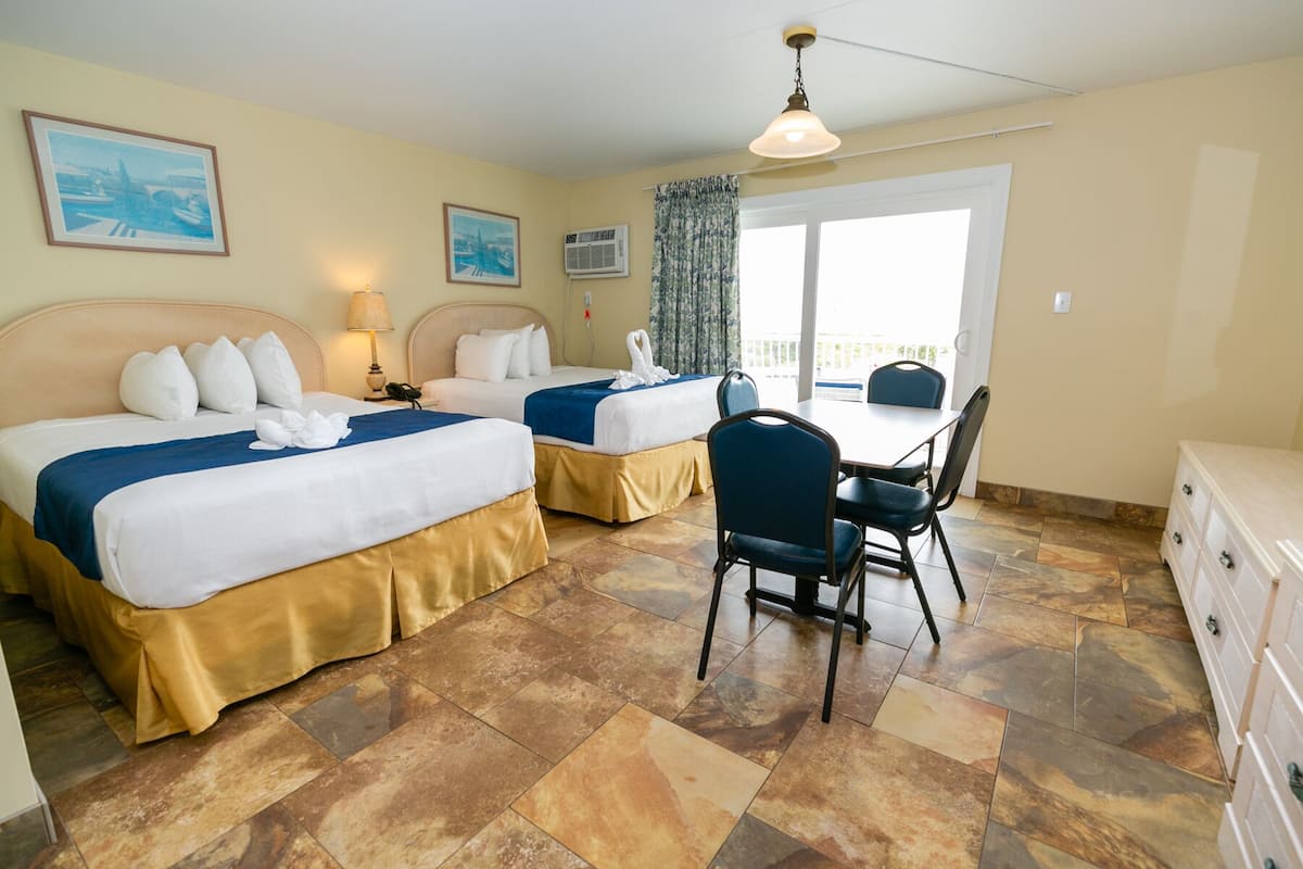 A hotel room with two beds, a dining table with chairs, a ceiling fan, and a window with curtains.