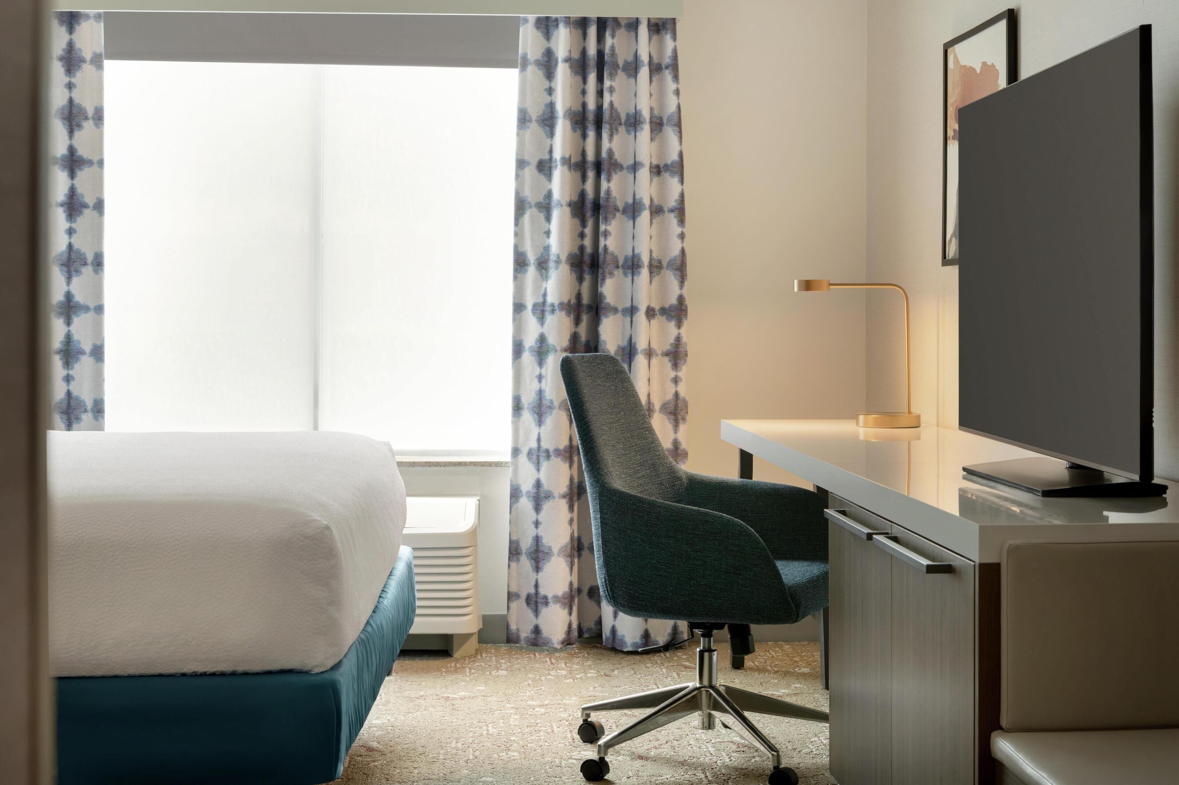 A hotel room with a bed, desk, chair, TV, and curtains.