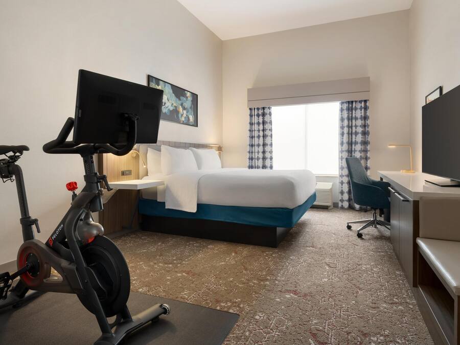 Habitación de hotel con cama, escritorio, silla, bicicleta estática, televisión y obras de arte en la pared.