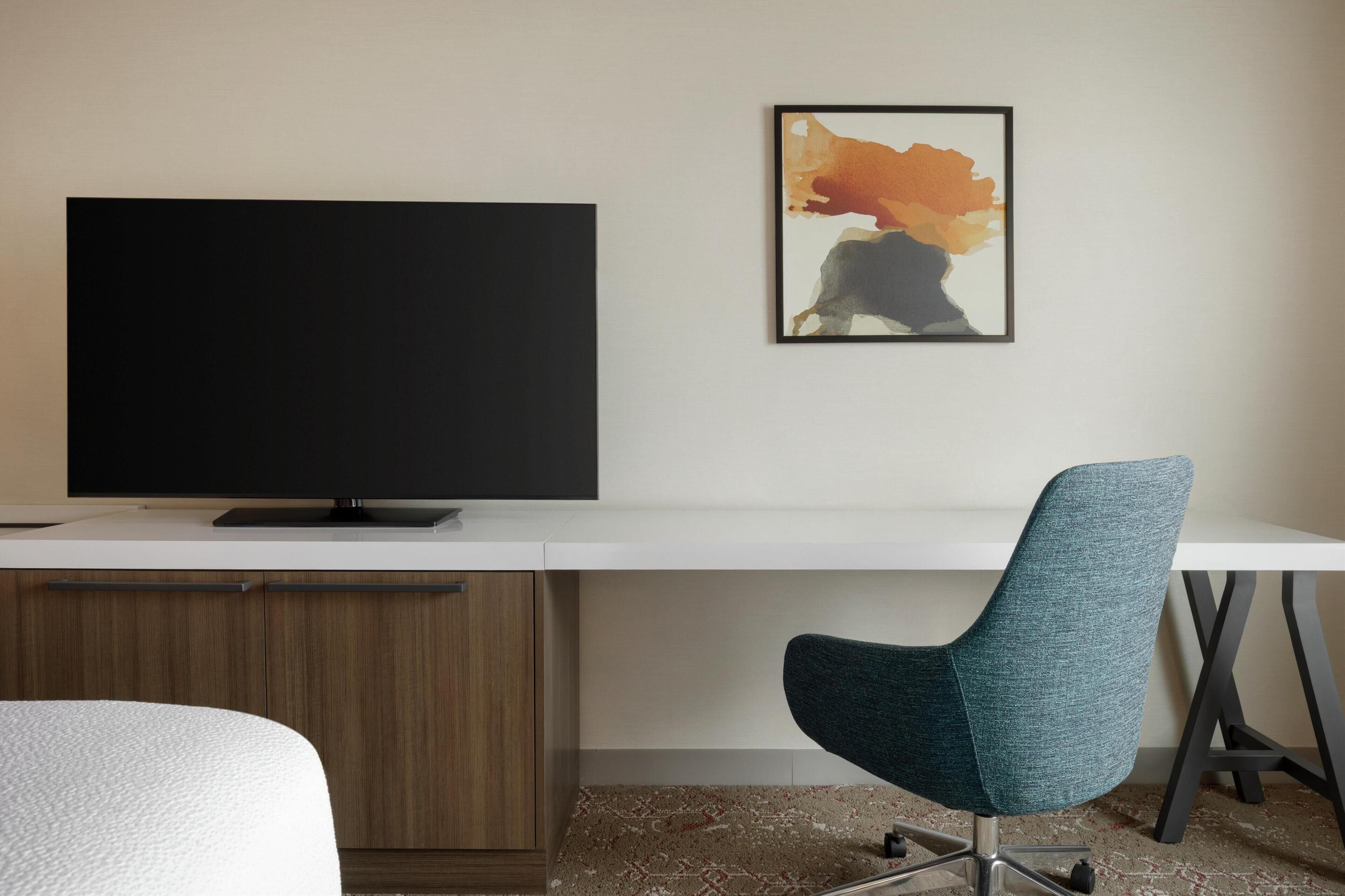Een moderne hotelkamer met een groot flatscreen-televisie, een bureau met stoel en een lijst met een abstract schilderij aan de muur.