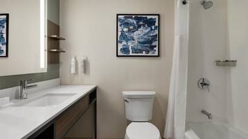 Une salle de bain moderne avec un lavabo, des toilettes et une baignoire, tous de couleur blanche. Un tableau abstrait encadré orne le mur, et il y a un rideau de douche.