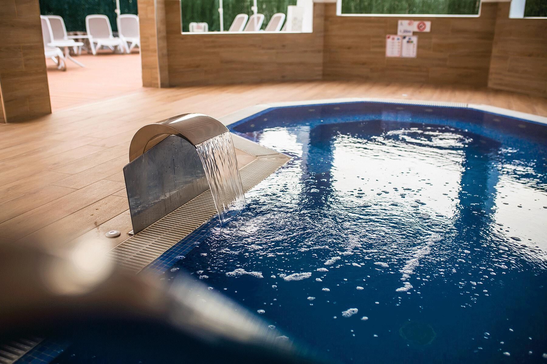 An indoor swimming pool with a water feature, wooden flooring, and lounge chairs in the background.