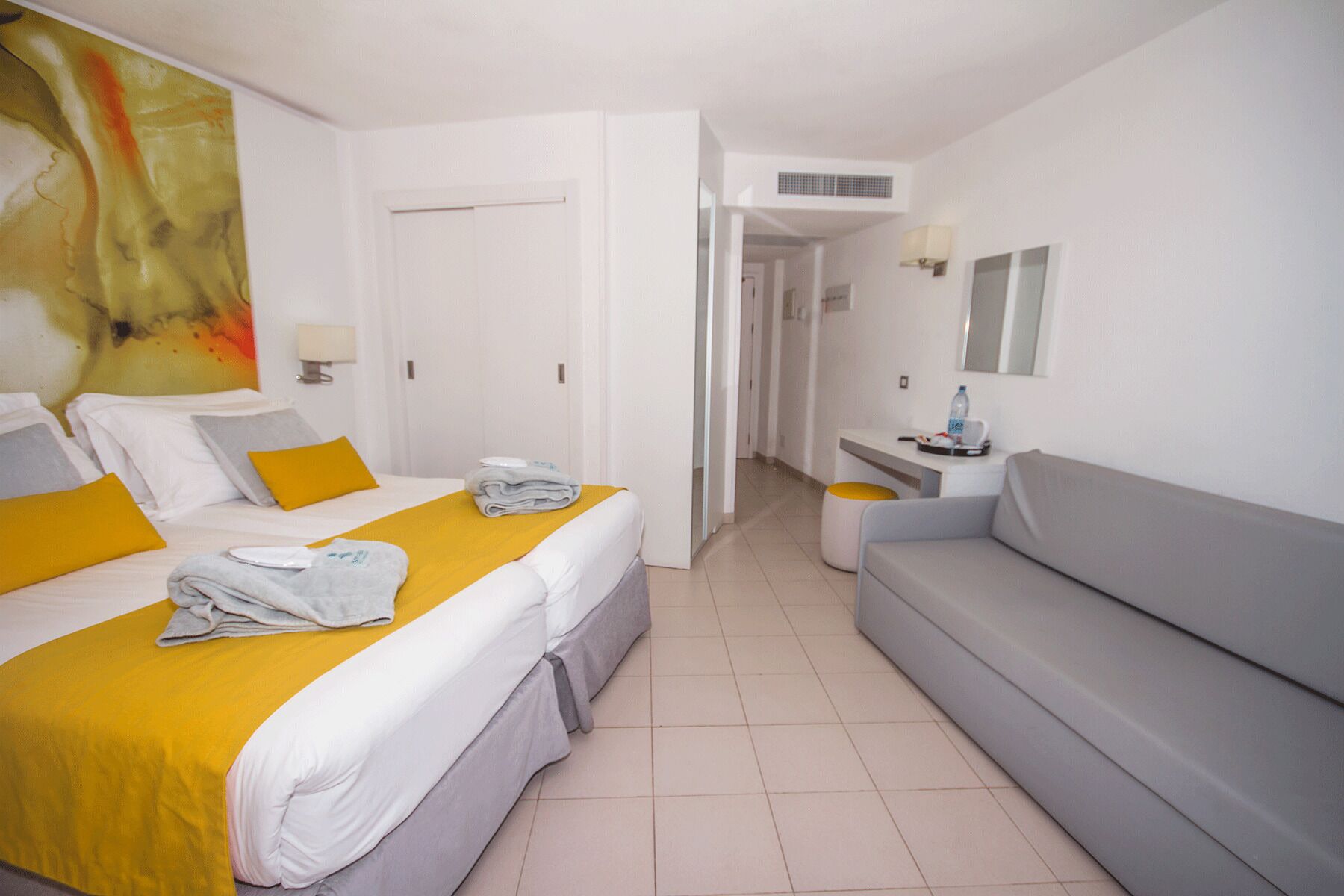 Habitación de hotel con dos camas, un sofá, un escritorio con lámpara y un cuadro abstracto grande en la pared.