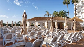 Een zwembadgedeelte met witte ligbedden en beige parasols, omringd door palmbomen en een wit gebouw met een betegeld dak.