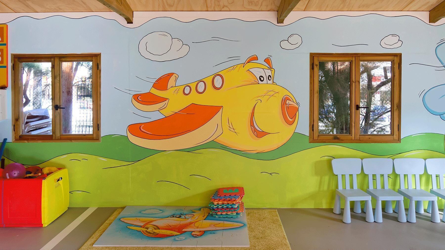 Sala de juegos para niños con un gran mural de un pez sonriente, un tapete de juego y sillas blancas.