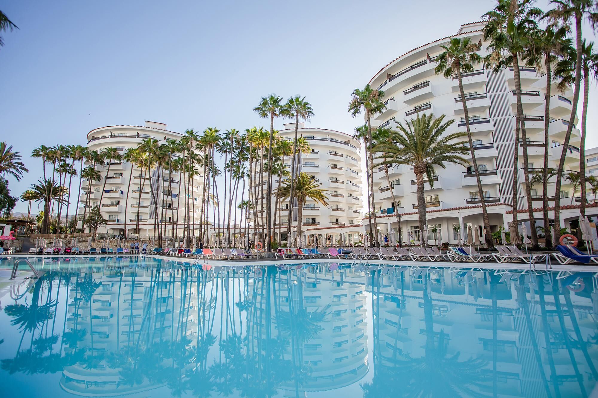 A resort pool area with multiple lounge chairs and umbrellas, surrounded by tall palm trees and a multi-story hotel building.