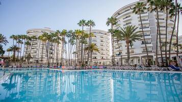 A resort pool area with multiple lounge chairs and umbrellas, surrounded by tall palm trees and a multi-story hotel building.