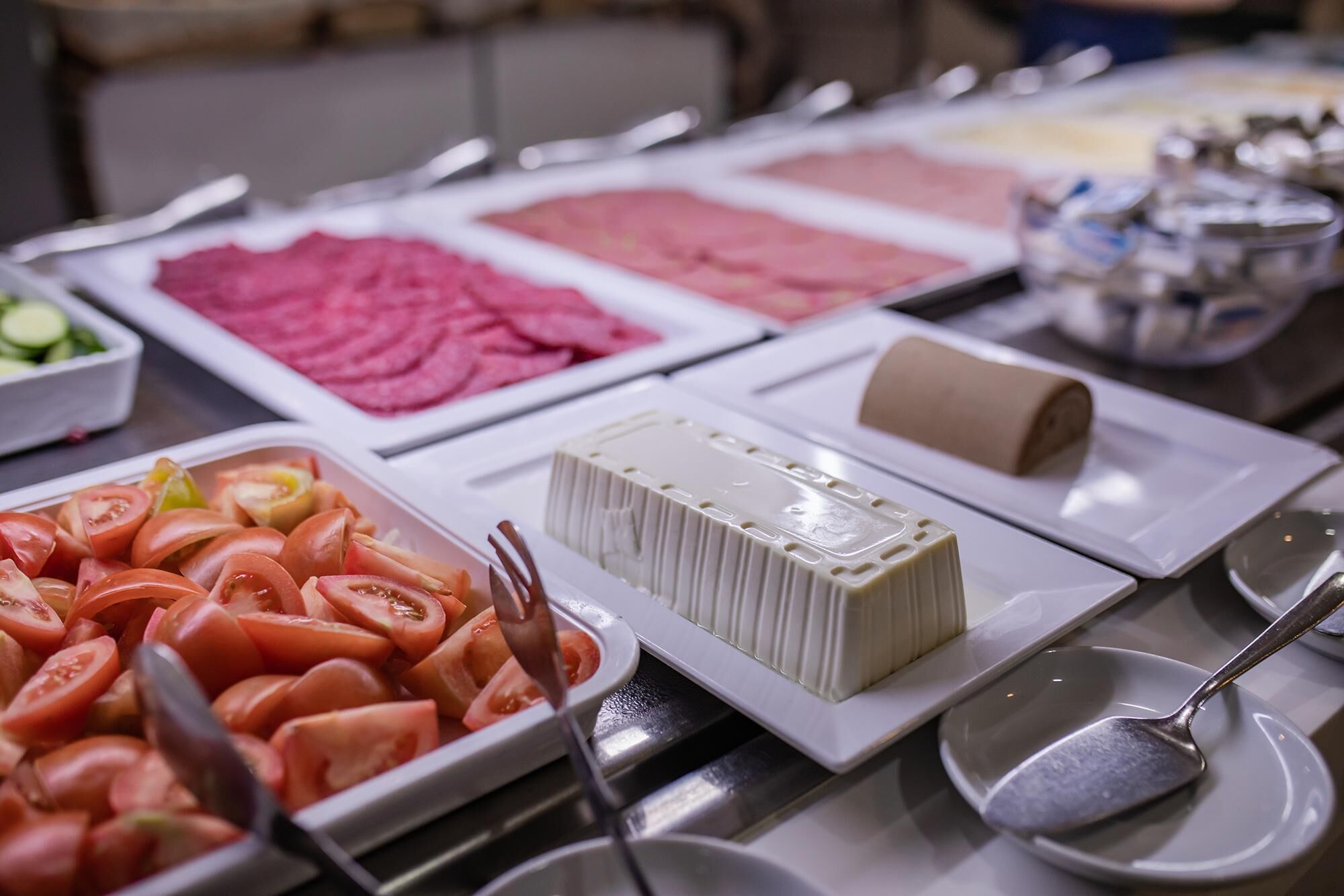Een buffet met diverse gerechten, waaronder plakjes tomaat, roze vlees en een blok tofu.