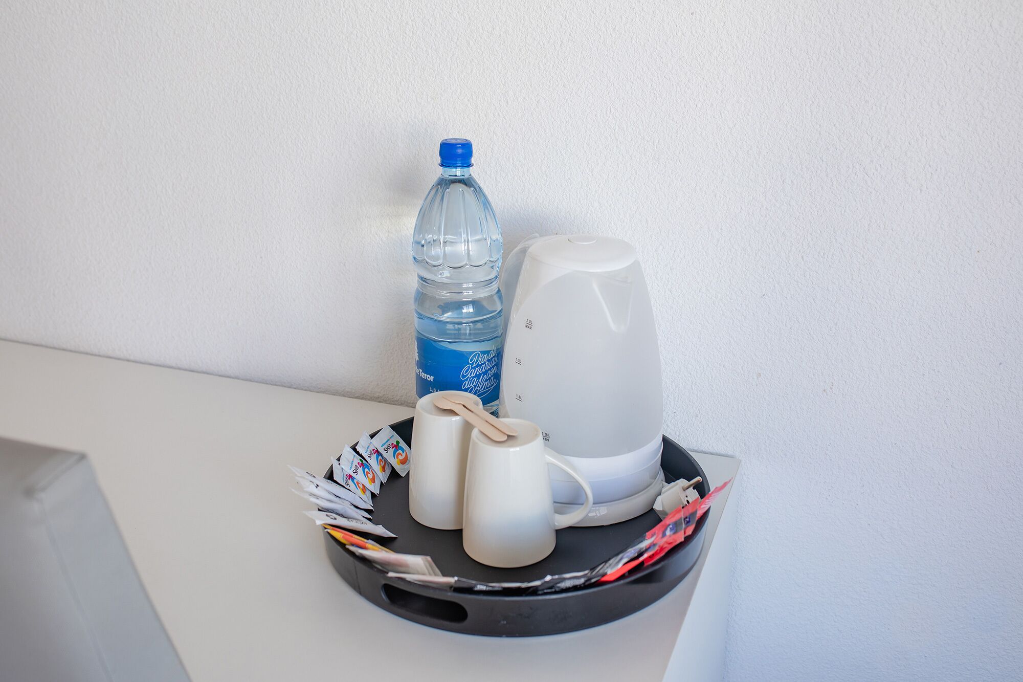 Ett litet bord med en vattenflaska, en vit mugg, en vit kopp och en vit vattenkokare.