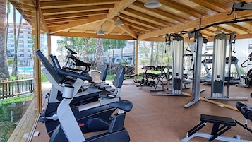 Et overdækket fitnessområde med træningsudstyr, herunder løbebånd og styrkemaskiner, under et trætag med store vinduer.