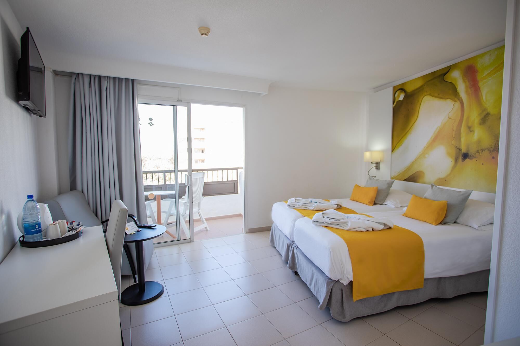 Habitación de hotel con dos camas, un escritorio con silla, televisor y balcón con vistas.