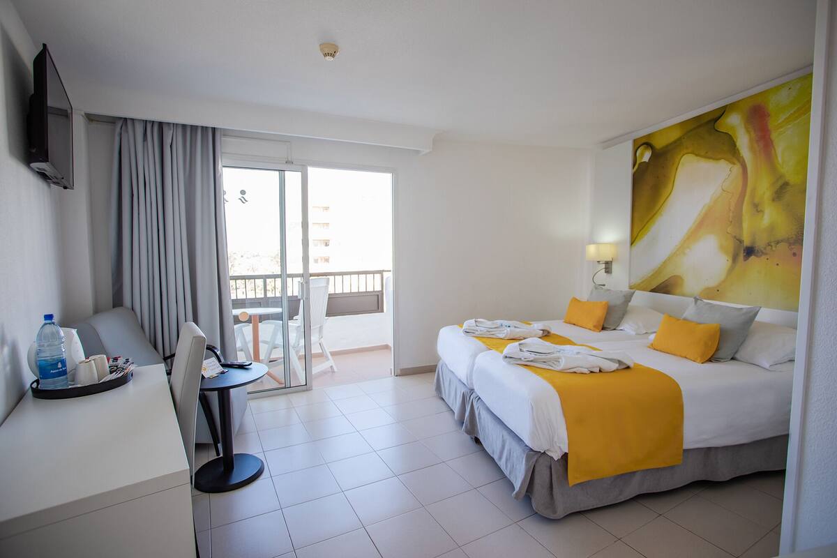 A hotel room with two beds, a desk, a chair, a TV, and a balcony with a view.