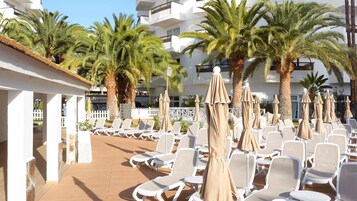 Zona junto a la piscina con sillas reclinables blancas y sombrillas color beige, rodeada de palmeras y un edificio blanco con balcones.