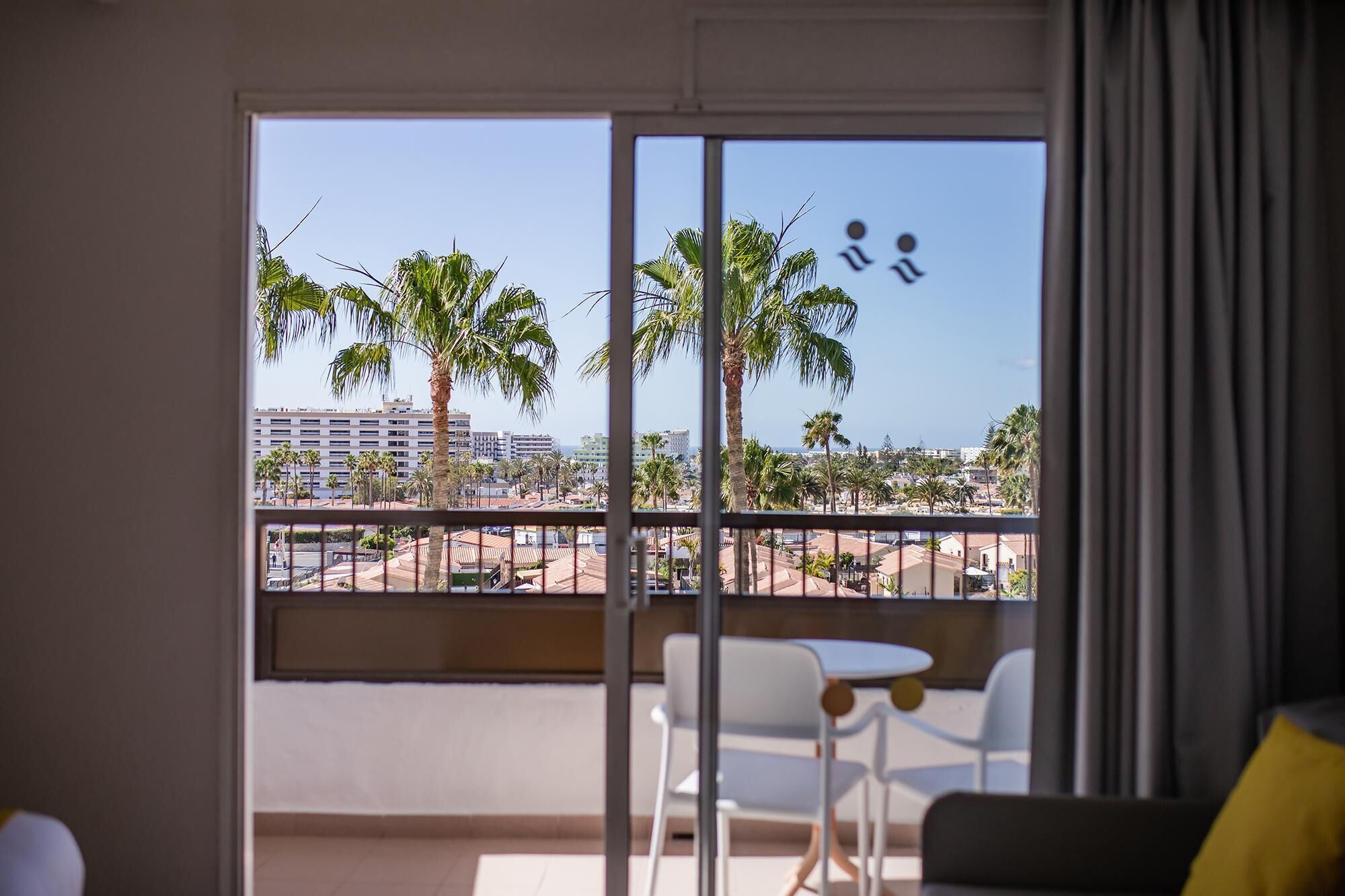 Un balcón con vistas a palmeras y edificios. Hay sillas blancas y una mesa en el balcón.