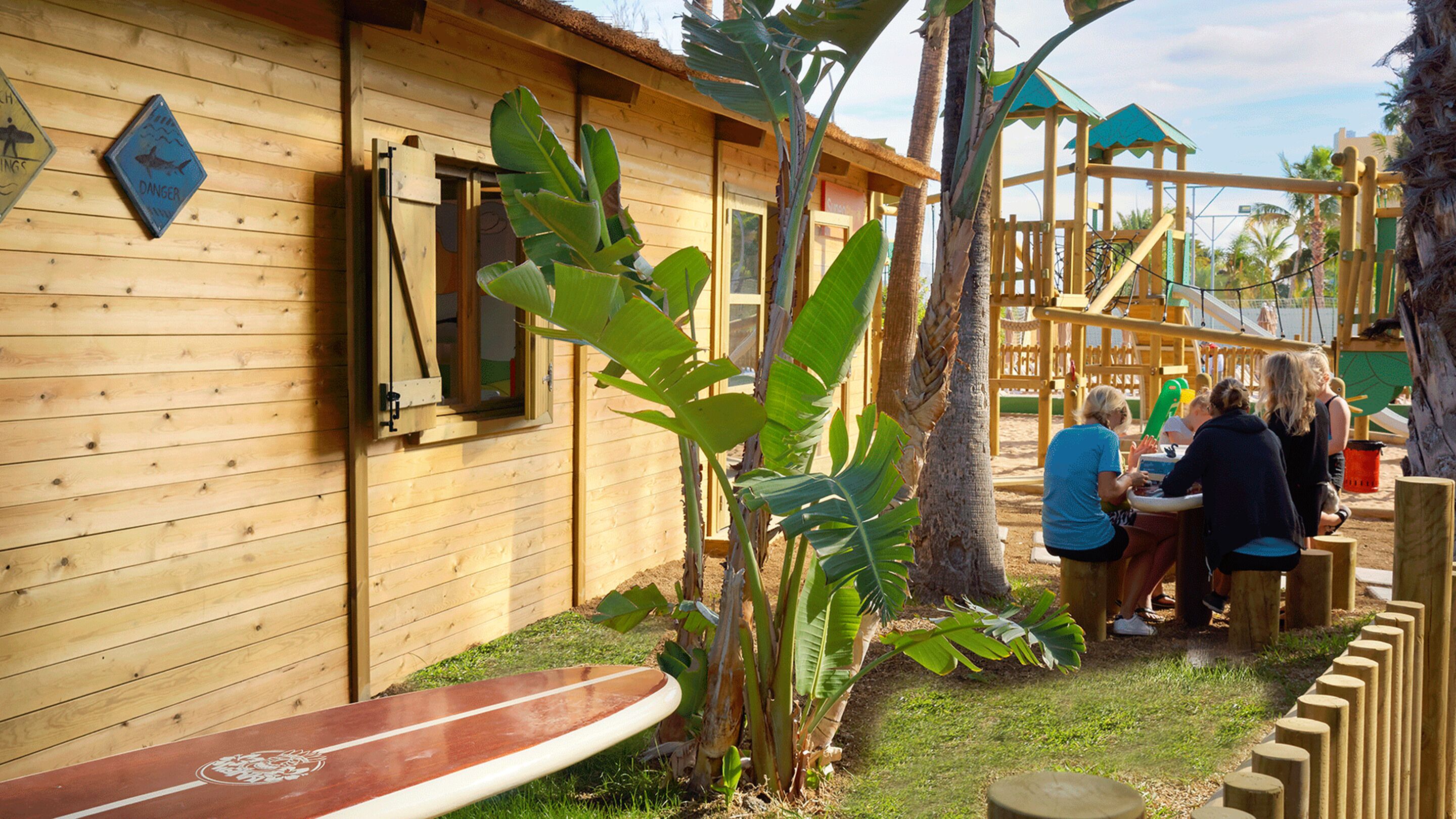 Una cabaña de madera con una ventana, una tabla de surf y una zona de juegos con un tobogán y estructuras para escalar.