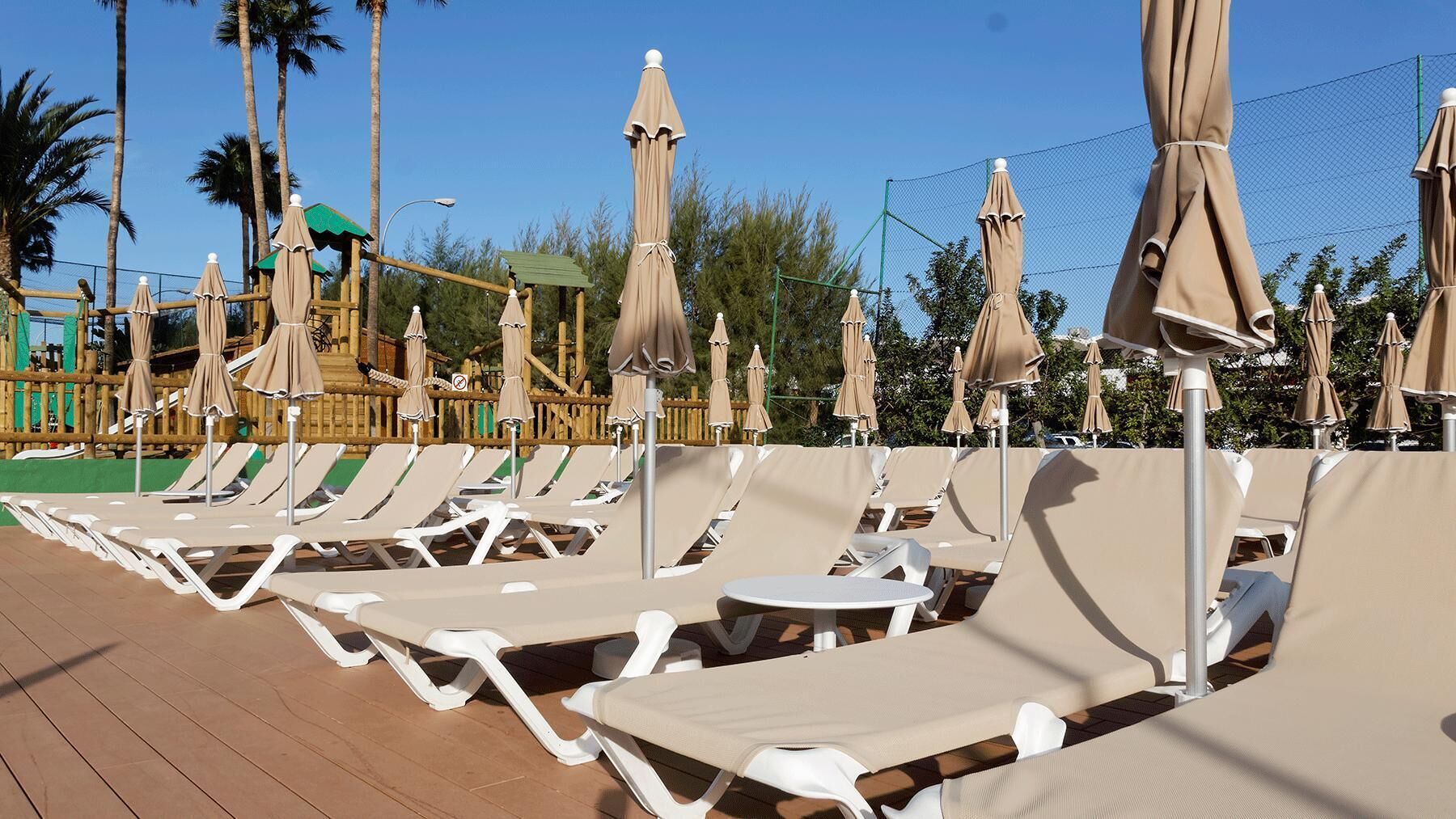 A poolside area with lounge chairs and umbrellas, a wooden deck, and a fence with trees in the background.