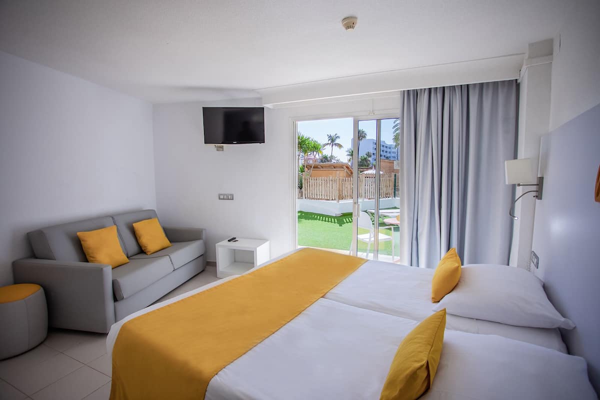 A hotel room with a bed, a sofa, a TV, and a balcony with a view of a pool and palm trees.