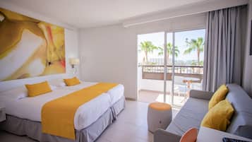 A hotel room with a bed, a sofa, a balcony with a view of palm trees, and a large abstract painting on the wall.