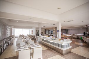 A modern hotel dining area with multiple tables and chairs, a buffet counter, and a kitchen in the background.