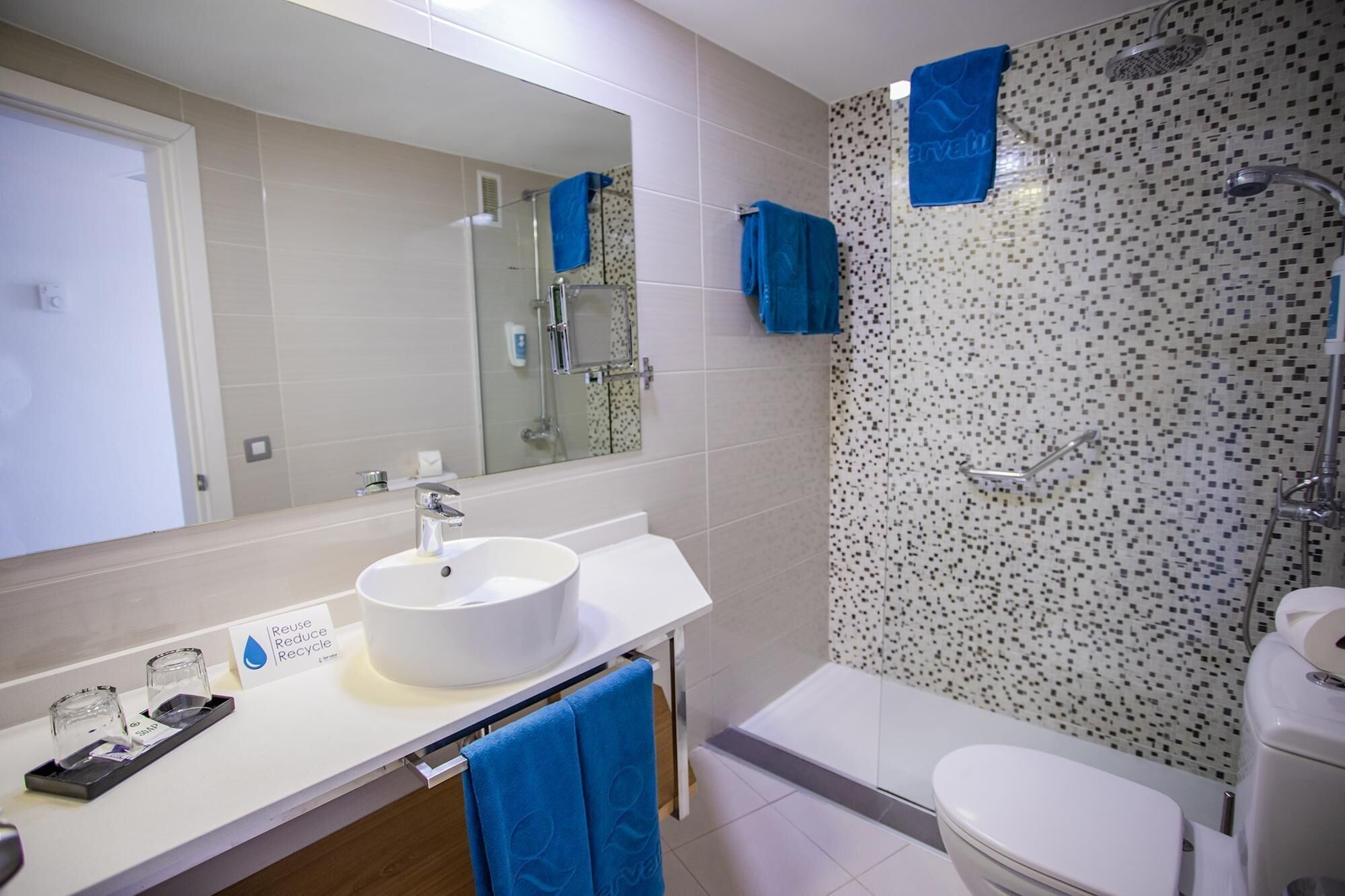 Un baño moderno con un lavabo blanco, un espejo, una ducha con un revestimiento de mosaicos y un inodoro.