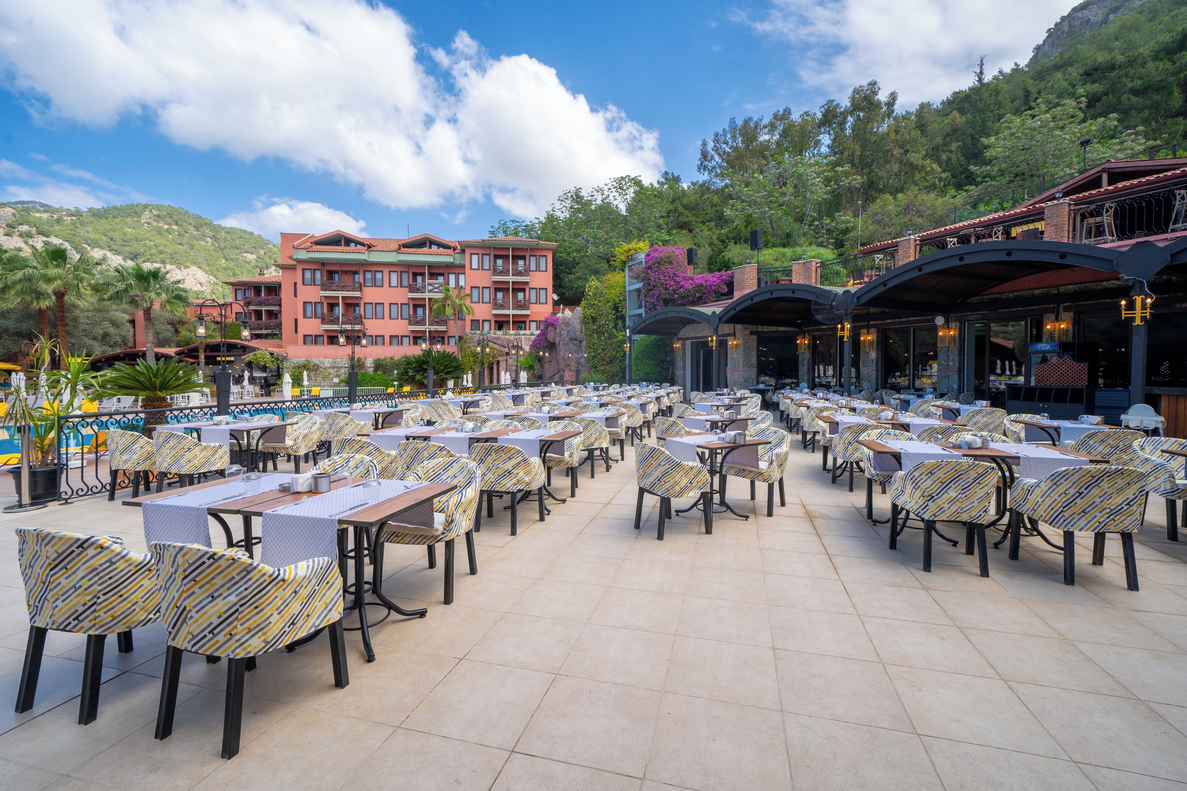 A spacious outdoor dining area with multiple tables and chairs, surrounded by lush greenery and a mountainous backdrop.