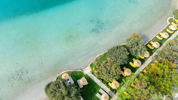 An aerial view of a resort with a clear beach, a row of yellow umbrellas, and lush greenery. - Sundia By Liberty Suncity - All inclusive (Fethiye)