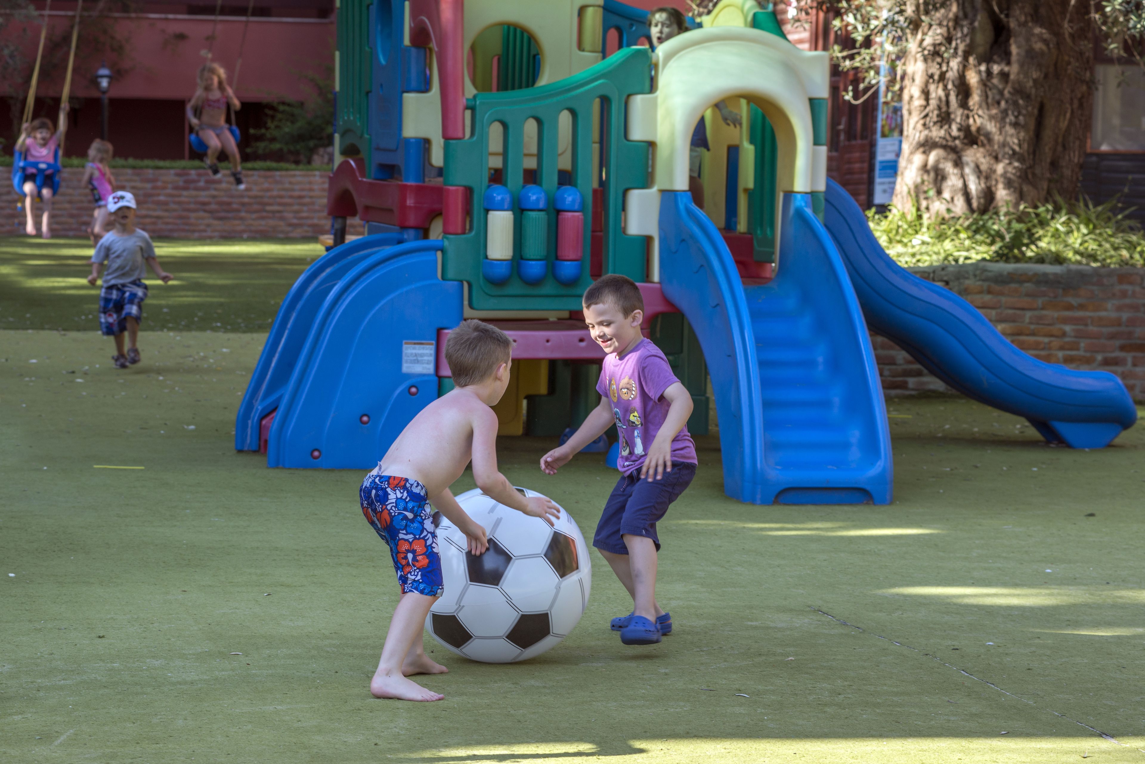 Des enfants jouent au ballon dans un parc avec des toboggans et des structures d’escalade.