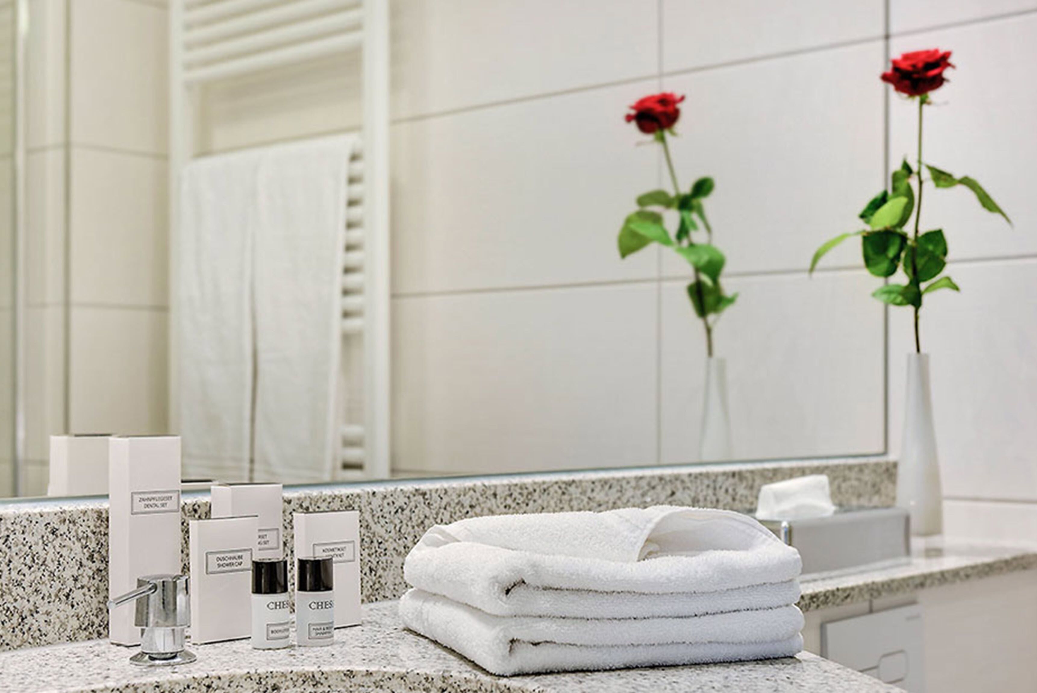A bathroom with a marble countertop, a stack of white towels, and two red roses in vases.