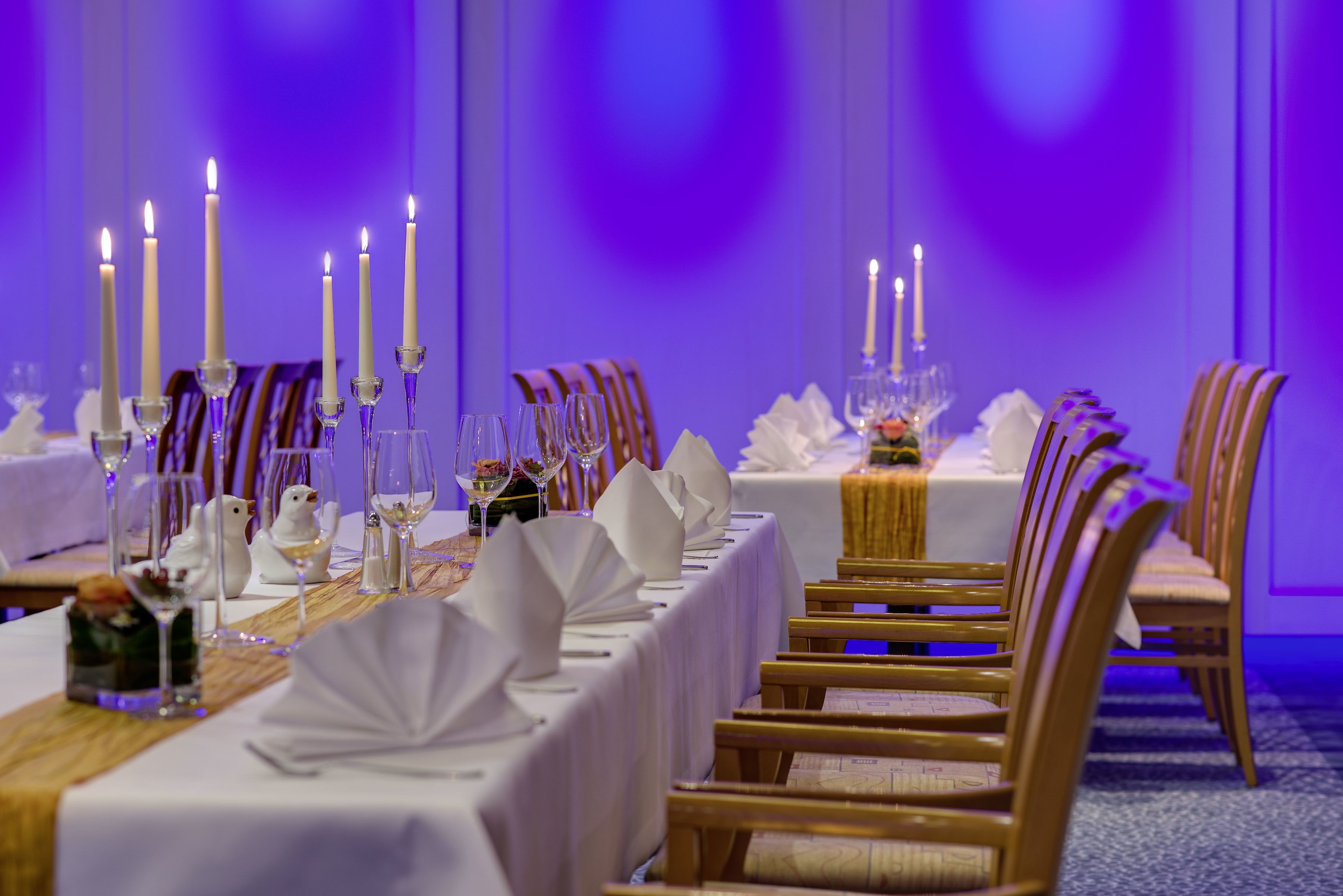 Une longue table à manger, recouverte de nappes blanches, agrémentée de bougies et de verres, dans une pièce éclairée d’une lumière violette.