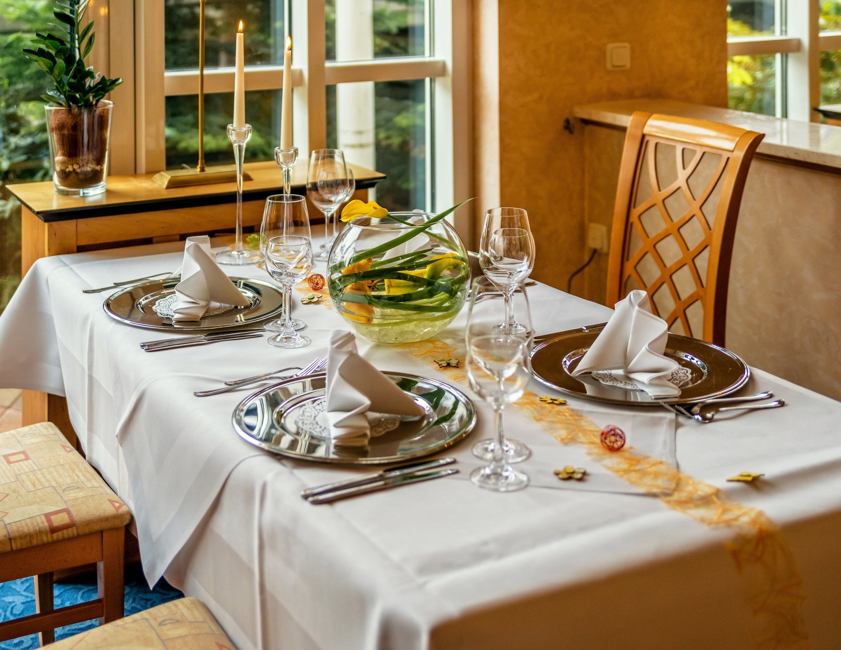 Una mesa de comedor preparada para cuatro personas, con manteles blancos, cubiertos y copas de vino.