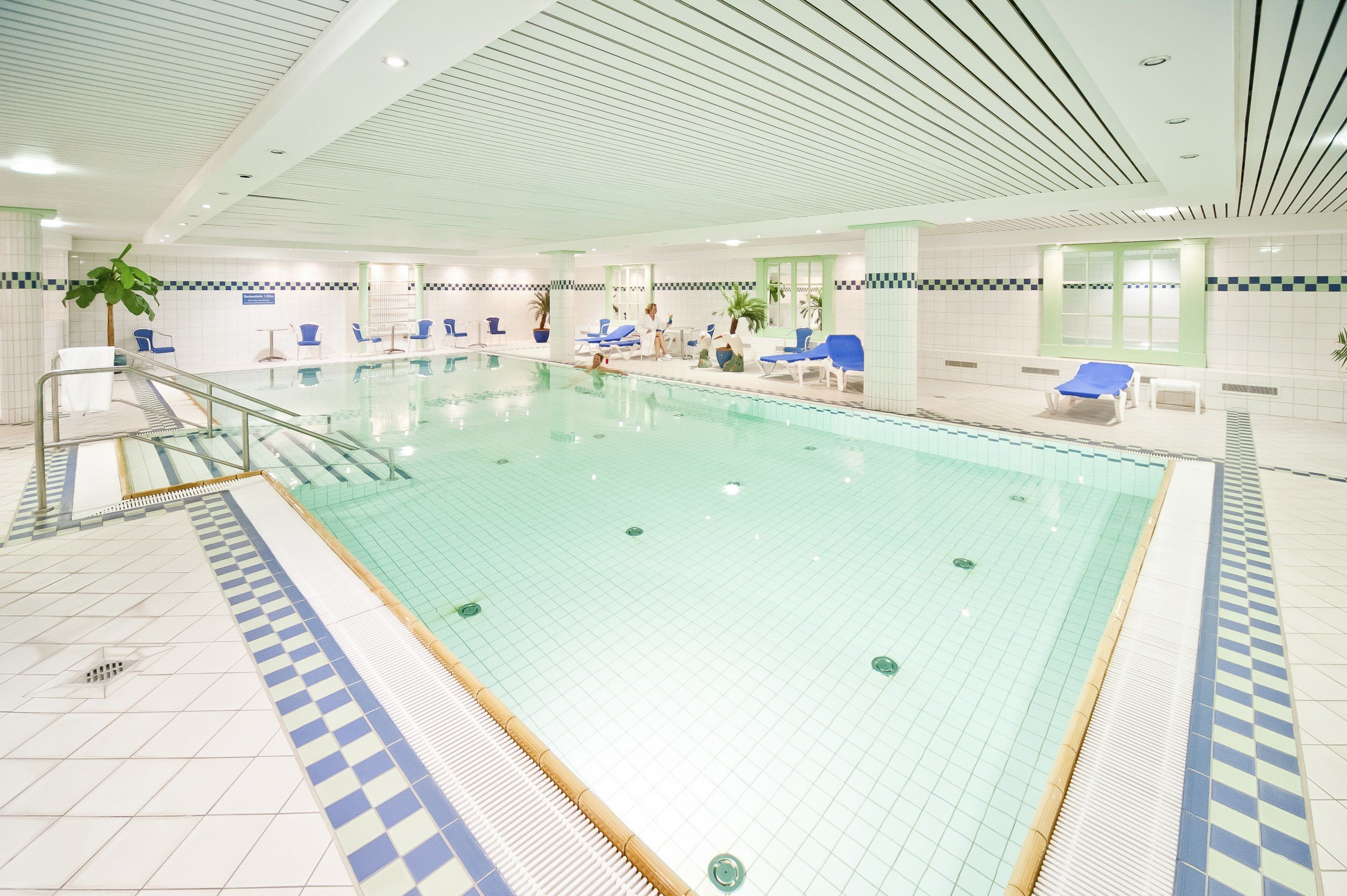 En stor inomhuspool med blå och vita kakelplattor på väggarna, solstolar och några få personer.