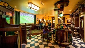 A bar area with a checkered floor, wooden furniture, and a large screen displaying a sports game.