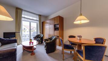 Ein Hotelzimmer mit einer Couch, einem kleinen runden Tisch, einem Esstisch mit Stühlen, einem Bücherregal und einem Fernseher.