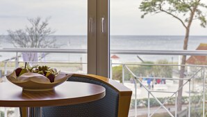 A table with a bowl of fruit and a chair, overlooking a balcony with a view of the sea and a tree. - Aquamarin Hotel & Spa (Kuehlungsborn)